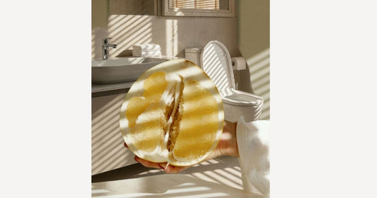 Close-up of a hand holding a halved yellow pomelo or grapefruit in a bright, modern bathroom with a toilet and sink in the background, featuring dramatic sunlight shadows from window blinds.