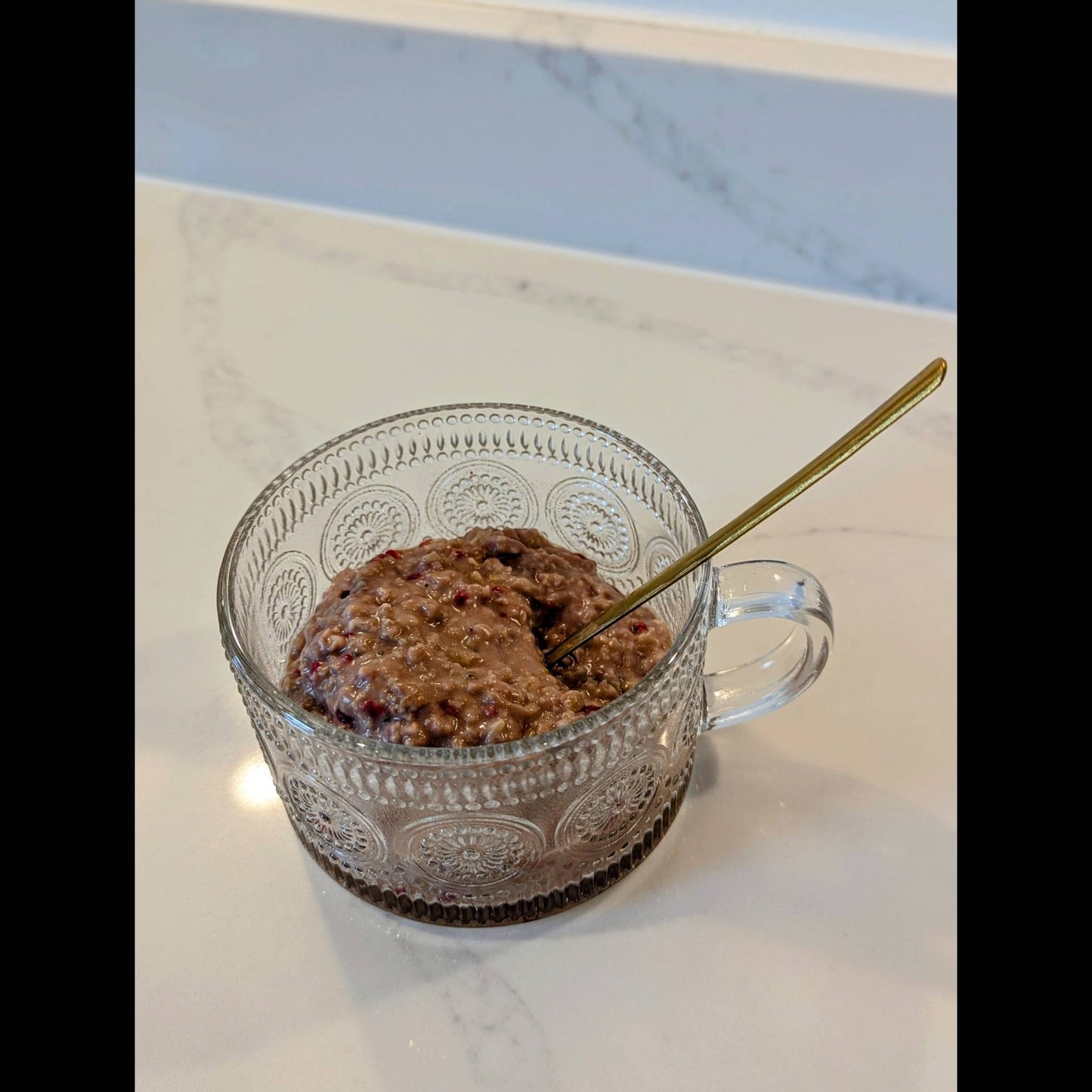 Clear glass mug with raspberry and cacao low FODMAP porridge and a gold spoon on a marble surface