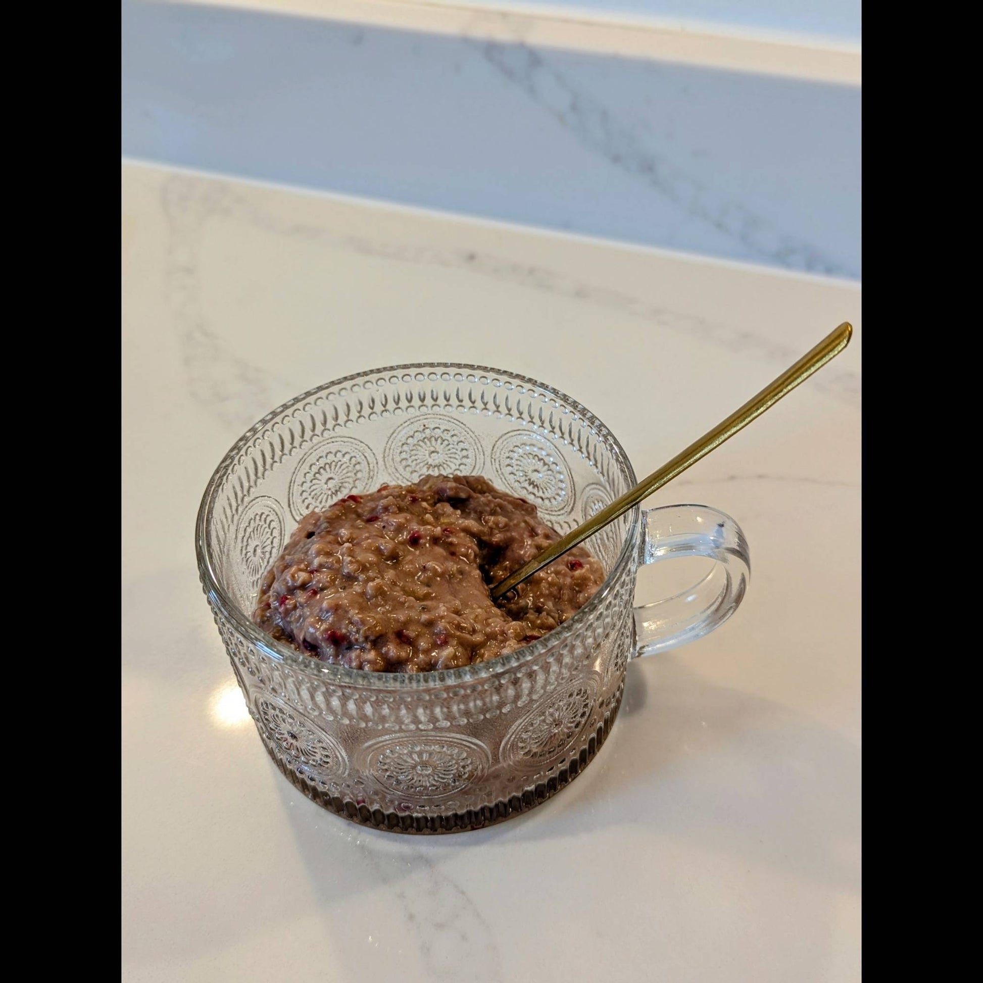 Clear glass mug with raspberry and cacao low FODMAP porridge and a gold spoon on a marble surface