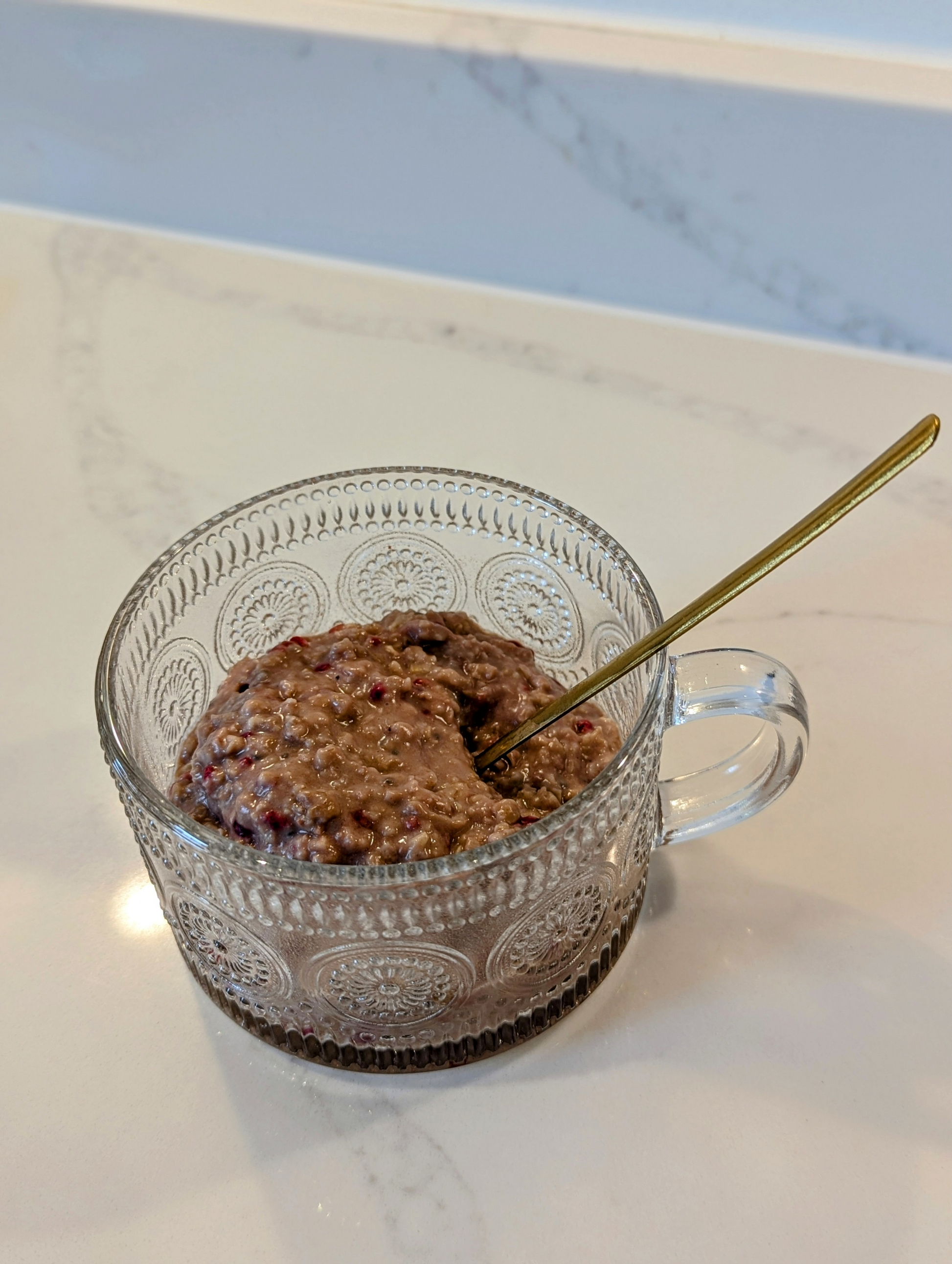Clear glass mug with raspberry and cacao low FODMAP porridge and a gold spoon on a marble surface