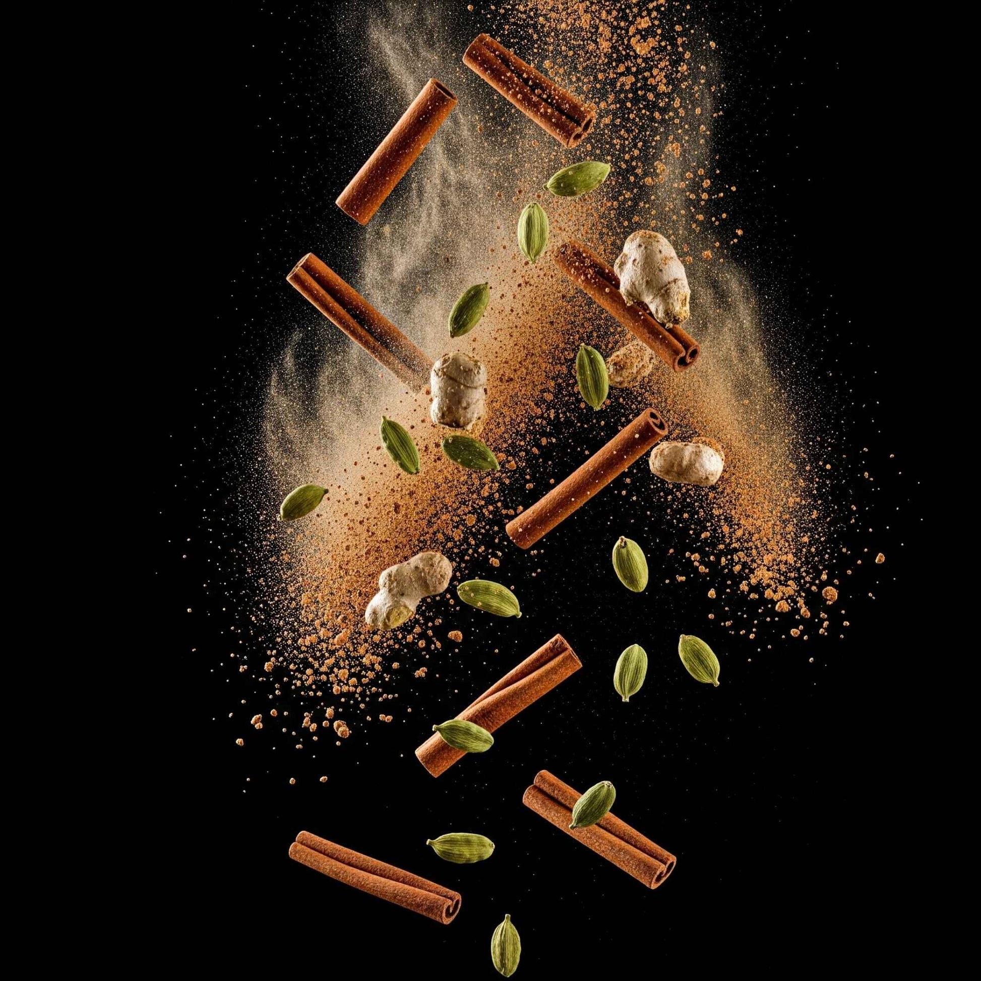Cinnamon sticks, cardamom pods and ginger pieces exploding in a cloud of spice on a black background.